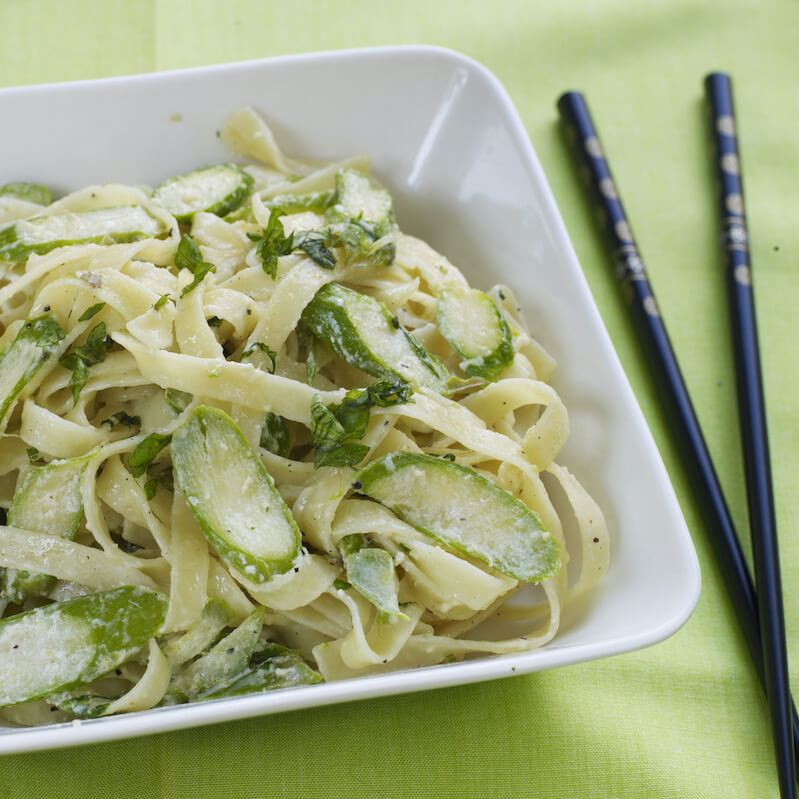 En firkantet skål med tagliatellepasta, asparges og ferske urter på en grønn duk.