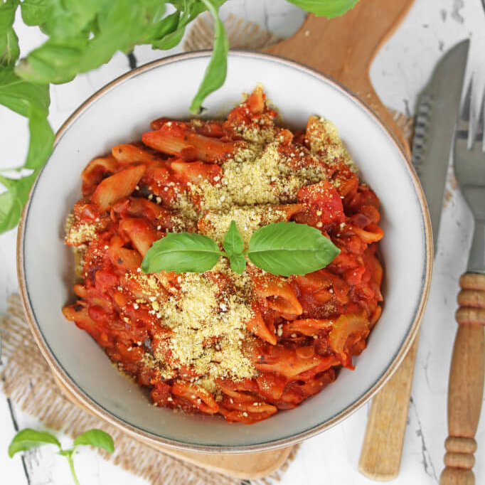 En skål pennepasta med tomatsaus og vegansk parmesan. Pyntet med fersk basilikumblad.