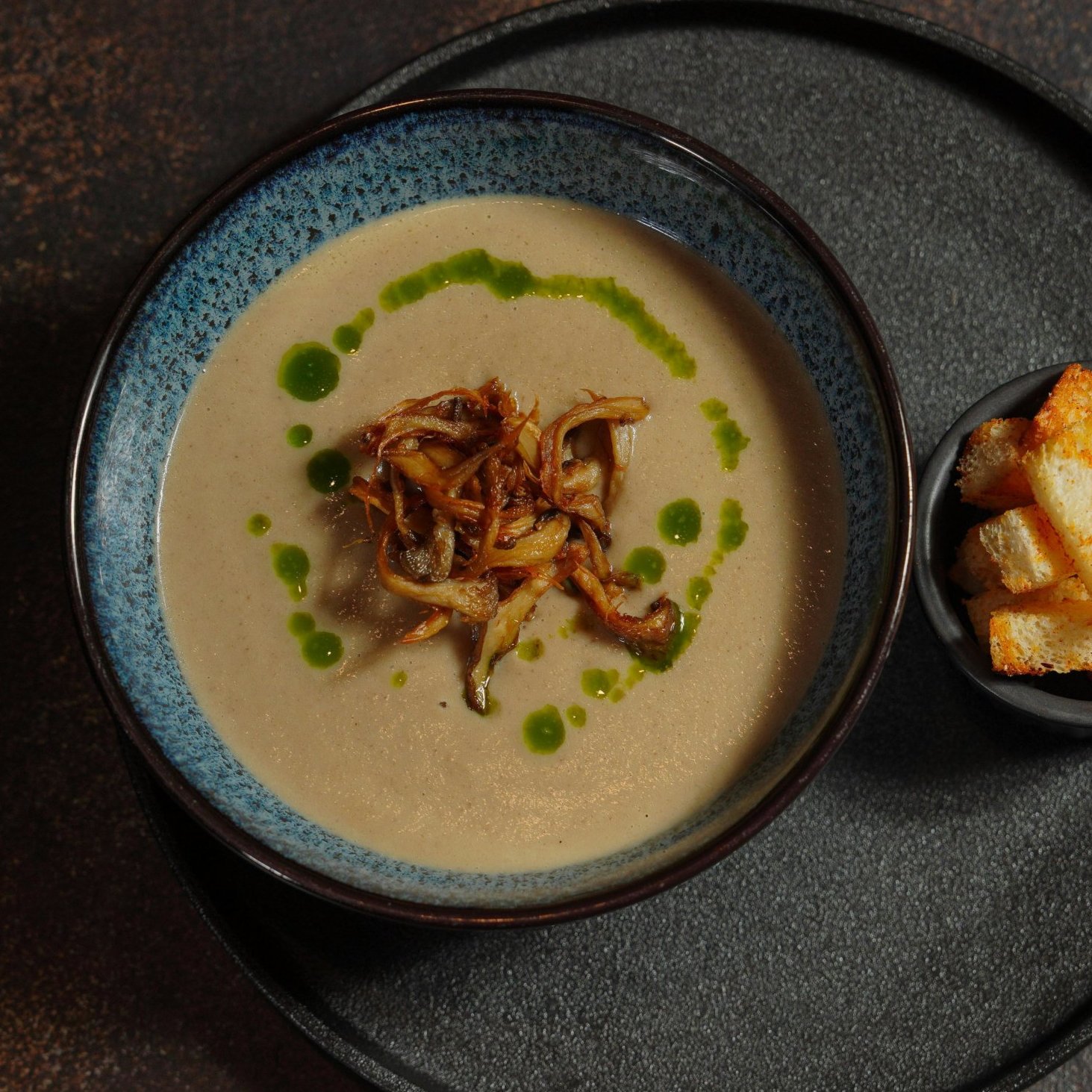 To skåler på et rundt serveringsbrett: èn med vegansk blomkålsuppe toppet md stekt sopp, og den andre fylt med krutonger.