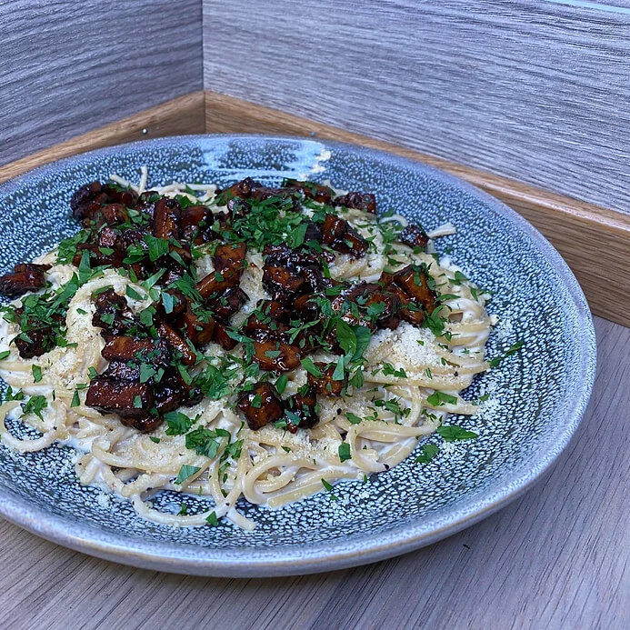 En blå tallerken med kremet vegansk pasta carbonara, toppet med friske urter og plantebasert bacon av tofu.