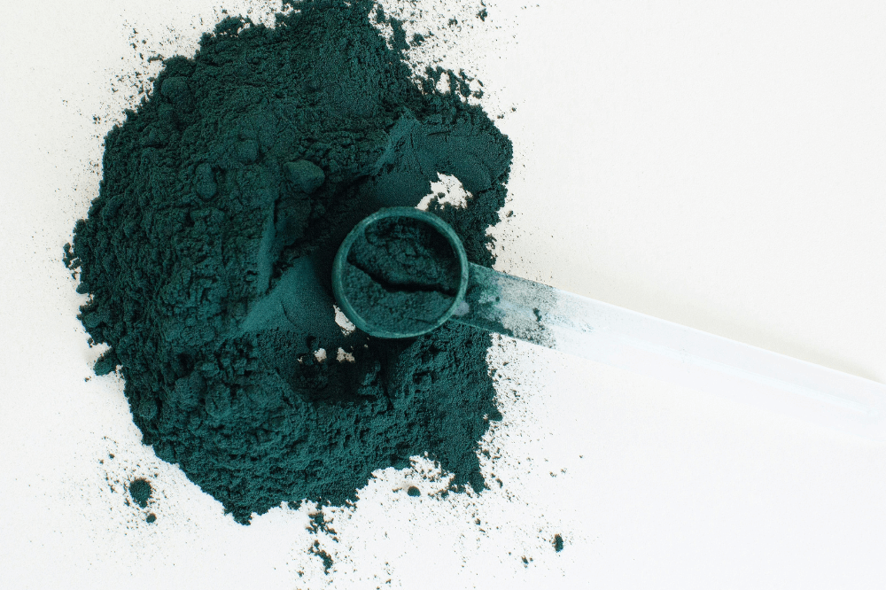 Blågrønt spirulinapulver spredt utover en hvit overflate med en måleskje