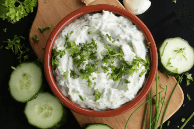 En terracottaskål fylt med plantebasert tzatziki, som har et dryss av friske urter på toppen. Skålen er på en trefjøl, or rundt er det agurkskiver gressløk og hvitløksbåter.