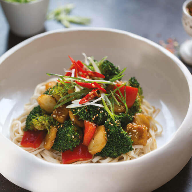 En hvit, dyp skål med tykke nudler, stekte grønnsaker og sorø tofu.