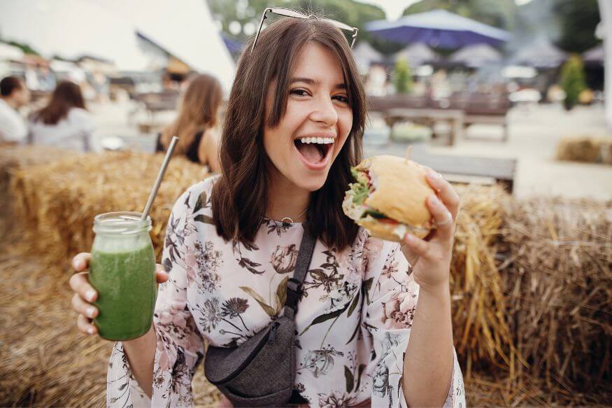 Smilende ung kvinner som spiser en vegetar-burger og drikker et glass med grønn juice eller smoothie.